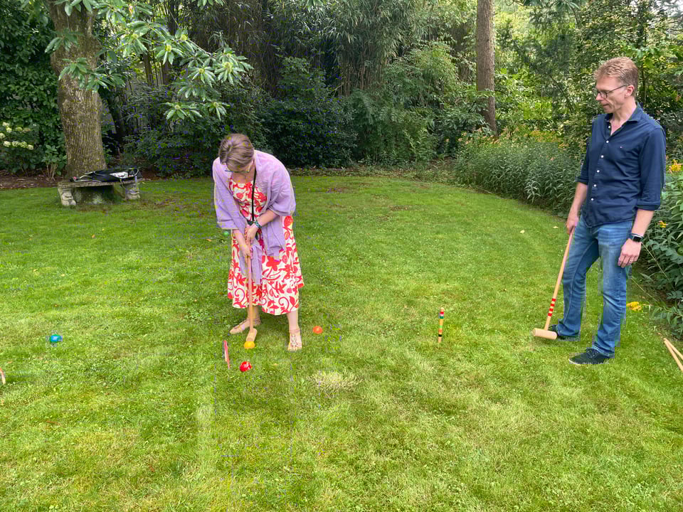 A woman plays croquet hitting a red ball while a man looks on.