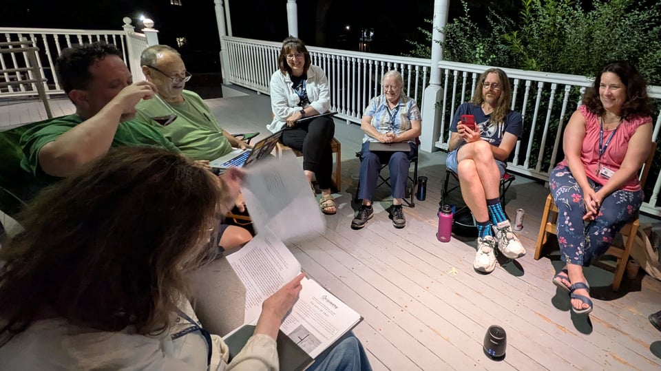 A bunch of writers sit in a circle on a huge porch. The camera is angled so you can see seven of them. There are drinks and papers and a laptop. Someone is preparing to read.