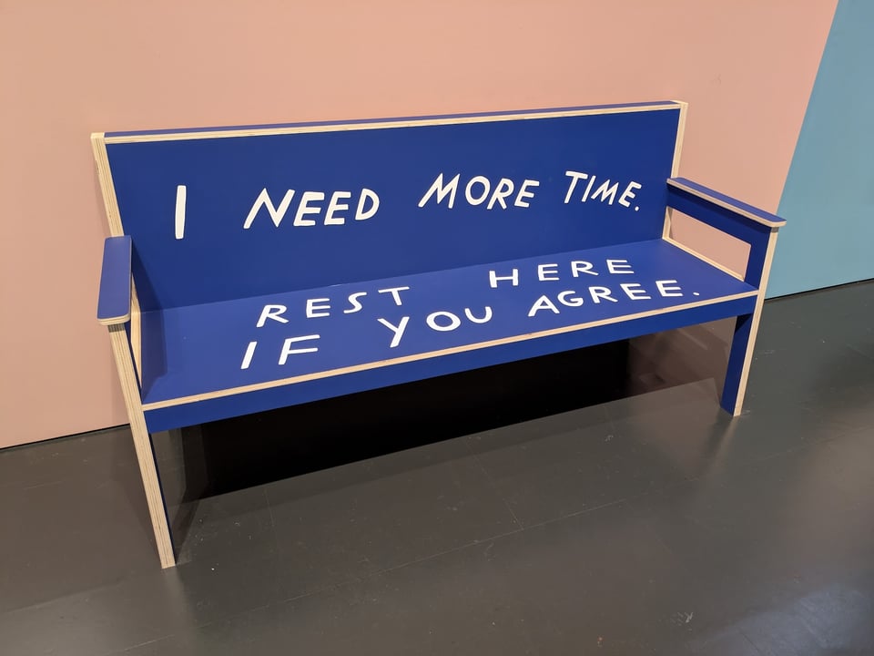 A long blue wood bench with armrests. Written on it in white text is "I need more time. Rest here if you agree."
