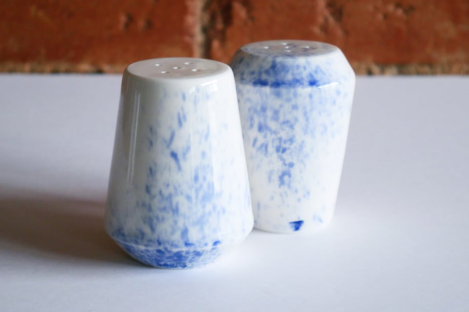 A set of two inverted angular salt and pepper shakers – one wide at the top, the other wide at the base – each with five holes, and a rubber stopper to close up the container. The porcelain gives it a bright white finish, with a clear gloss glaze, and a splash of blue cobalt wash.