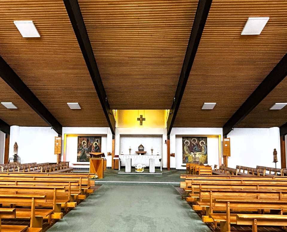 Looking up the short aisle of a wide church. There are rows of beech benches lined up on the floor. Overhead is a huge, wood-lined ceiling that is sloping down towards the alter. This sits between windows that cause it to glow golden.