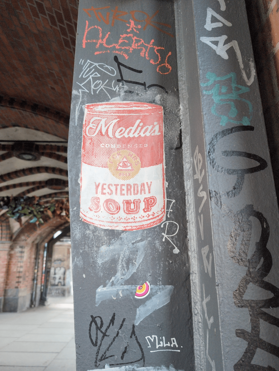 Graffiti on a metal support beam for a bridge: There are a variety of text tags, but the focus is a Campbell's Soup can that reads instead "Media's condensed yesterday soup," and the Campbell's logo is a pyramid with an eye in the middle with the text "FCK WRS 2013."