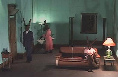 A green room with brown carpet and furniture. Three human-sized rabbits are arranged awkwardly around the room