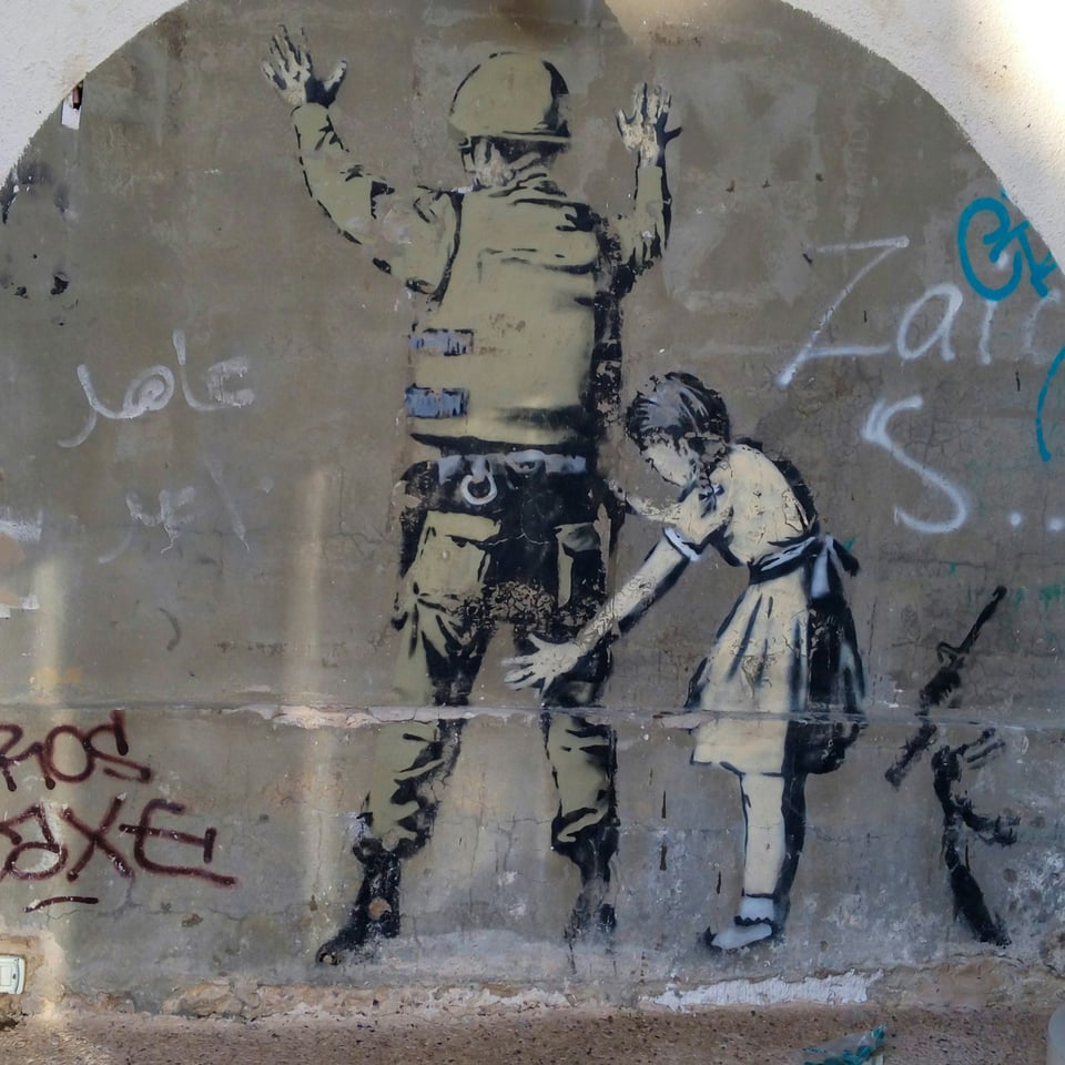 Graffiti art of a girl in a dress patting down a soldier in body armor and a helmet who has his hands up against the wall. She has broken his gun and it's discarded next to them.