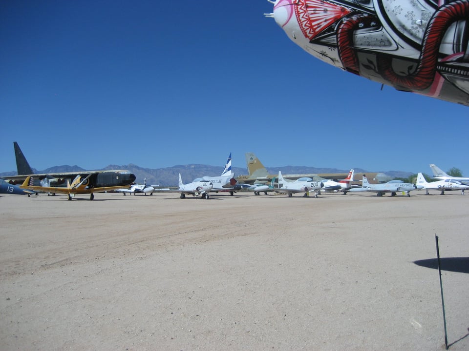 An open area of brown, sandy ground, on which is parked a variety of airplanes with mountains in the background.