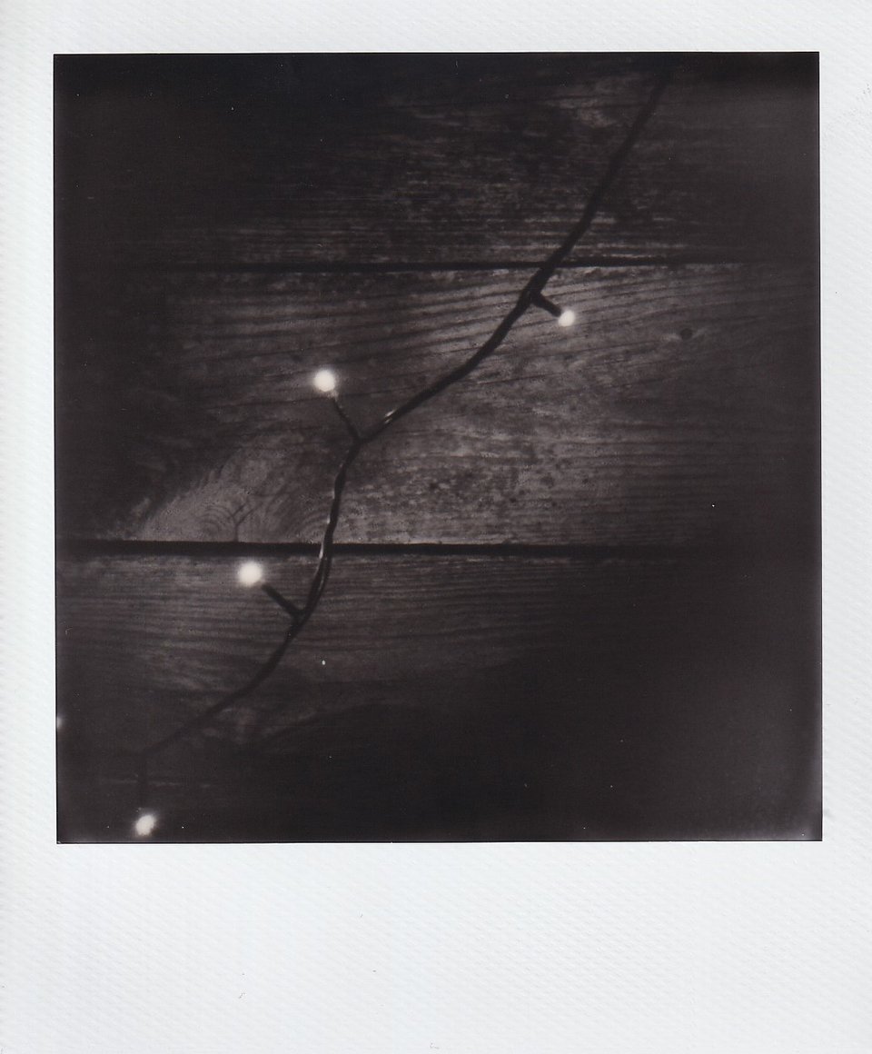 A black and white Polaroid of a string of white Christmas lights laid against a wood wall.