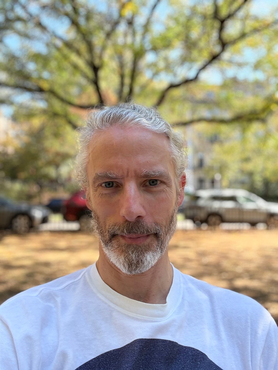 Selfie of white man with bear, wearing a white pullover with a big blue circle; there's a tree behind him and it looks like the branches are emanating from his head
