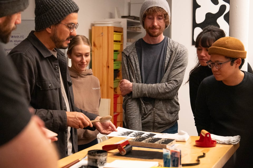 A group of people standing around a table watching Justin demonstrate block printing.
