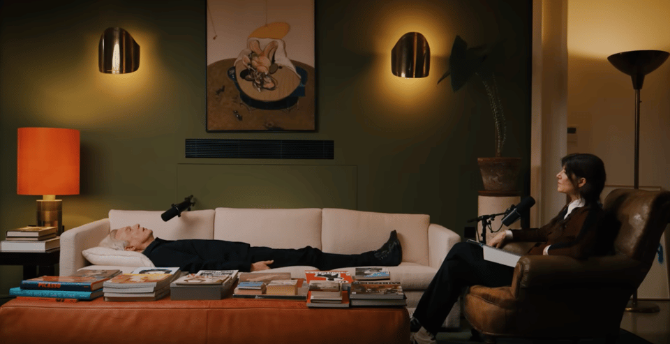 Screenshot from a podcast video. A man (David Byrne) is lying on a couch. There is a microphone near to his mouth. A woman (Bella Freud) is sitting in a chair to the right of the picture. She too is talking into a microphone.