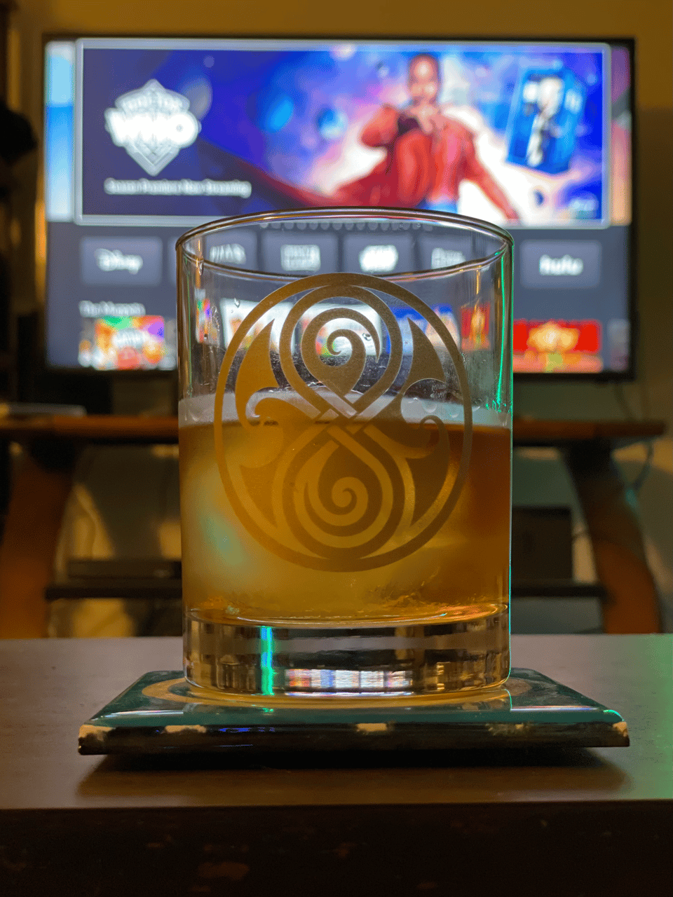 A glass of whiskey in a Time Lord cup in front of a glowing screen featuring the 15th Doctor