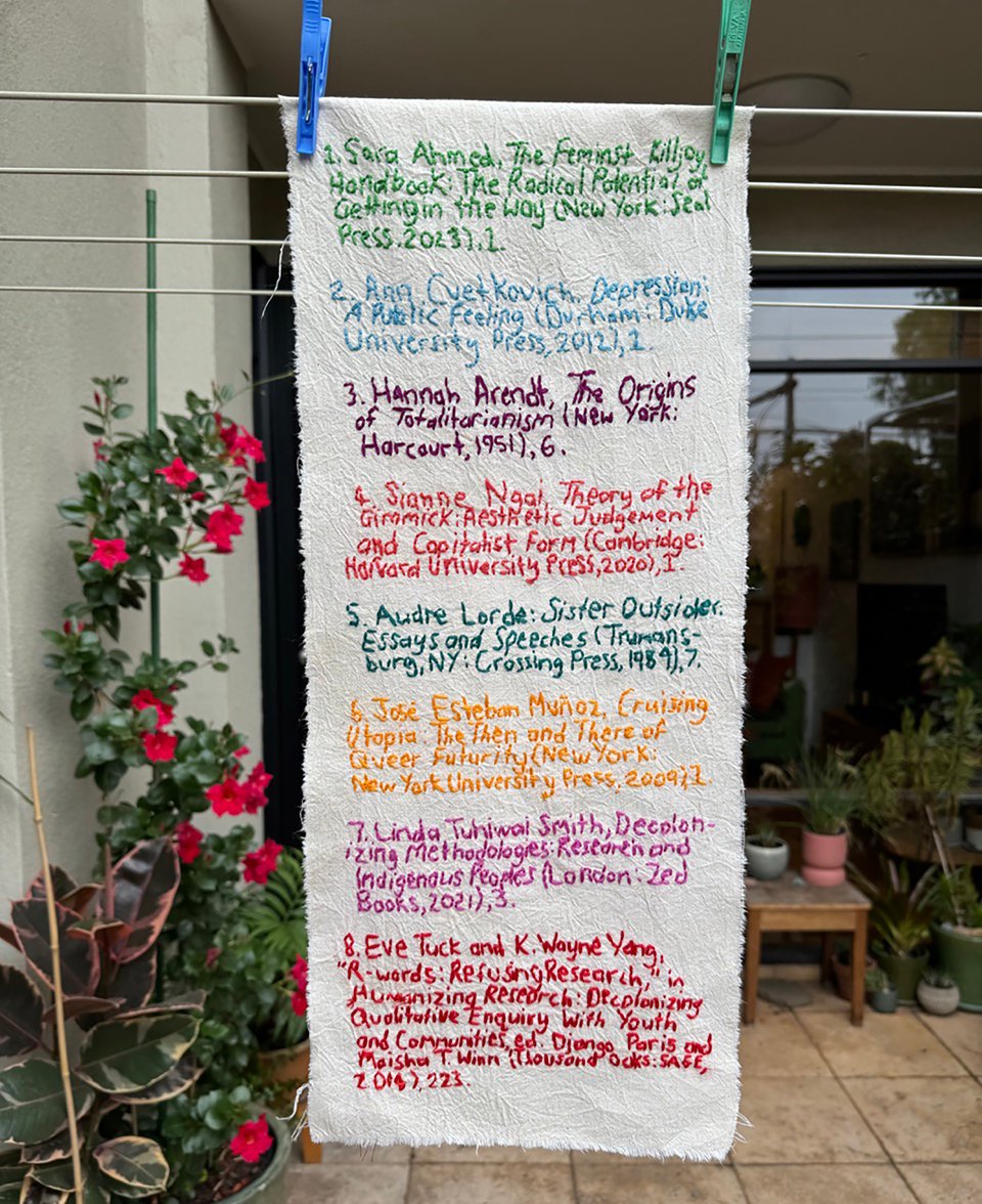 A piece of calico hangs from a domestic courtyard clothesline. Embroidered in different coloured threads on the calico is a list of 8 footnotes from queer, feminist, and First Nation scholars.