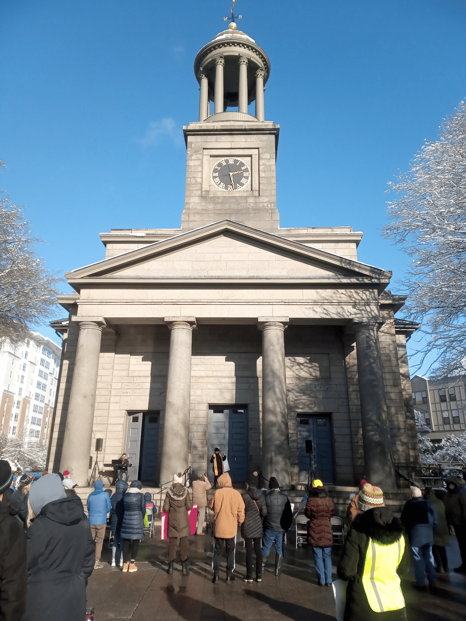 On a sunny day in January a small crowd gathers in front of the United First Parish Church, which is a grey neoclassical building with pillars and a bell tower.
