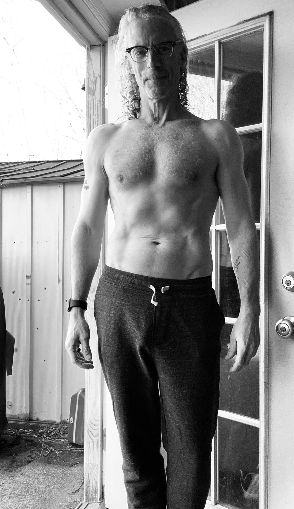 B/w photo of slim, shirtless white man in sweatpants, standing indoors by an open door. He's wearing glasses and has long wet hair. His body looks okay. b/w photo of shirtless white man in sweatpants, standing indoors by an open door. He's wearing glasses and has long wet hair. His body looks pretty good, esp. since he's in his mid-50s