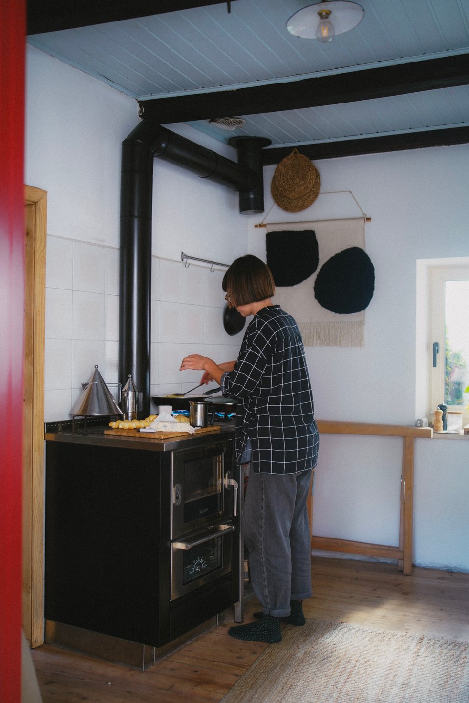[Elena in a minimal but beautiful kitchen with a stove, coffee pot, hanging textiles, window, wood floor, and jute rug,]