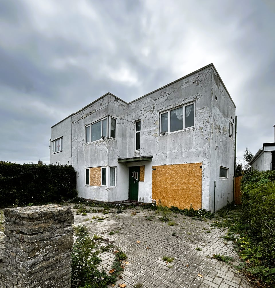 A 1930s house, like the others already described except the windows are modern uPVC frames. The ground floor is entirely boarded up with chipboard except, surprisingly, for the still original front door. The white render is filthy and starting to fail.