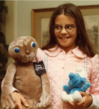 A girl in a bathrobe and glasses holding two stuffed animals: ET and a smurf