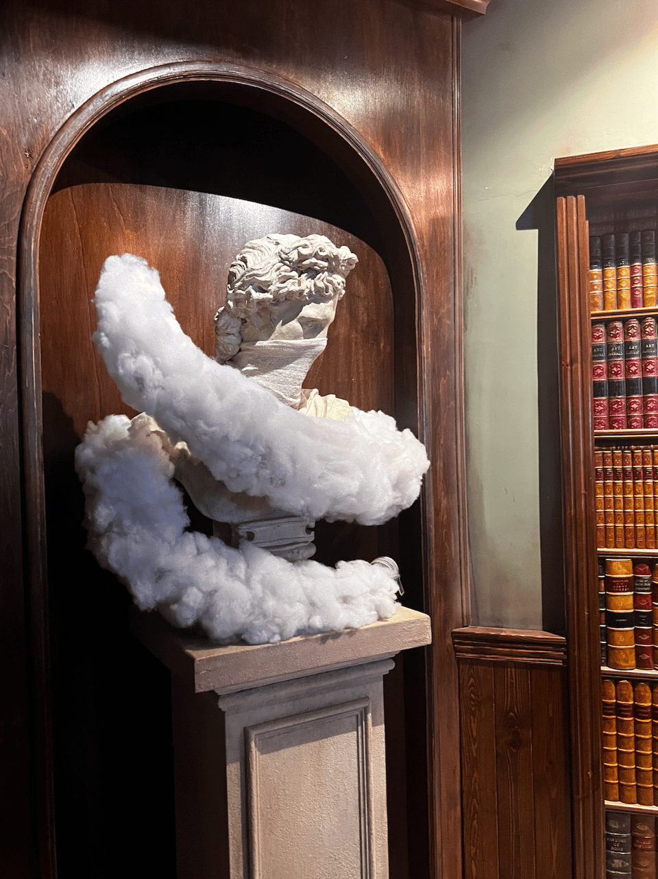 Photograph of a classical marble bust of a man with wavy hair, displayed inside a dark-wood arched niche. Wood paneling and leather-bound books line adjacent shelving. The man is wearing a mask, and a white cloud-like form surrounds the bust.