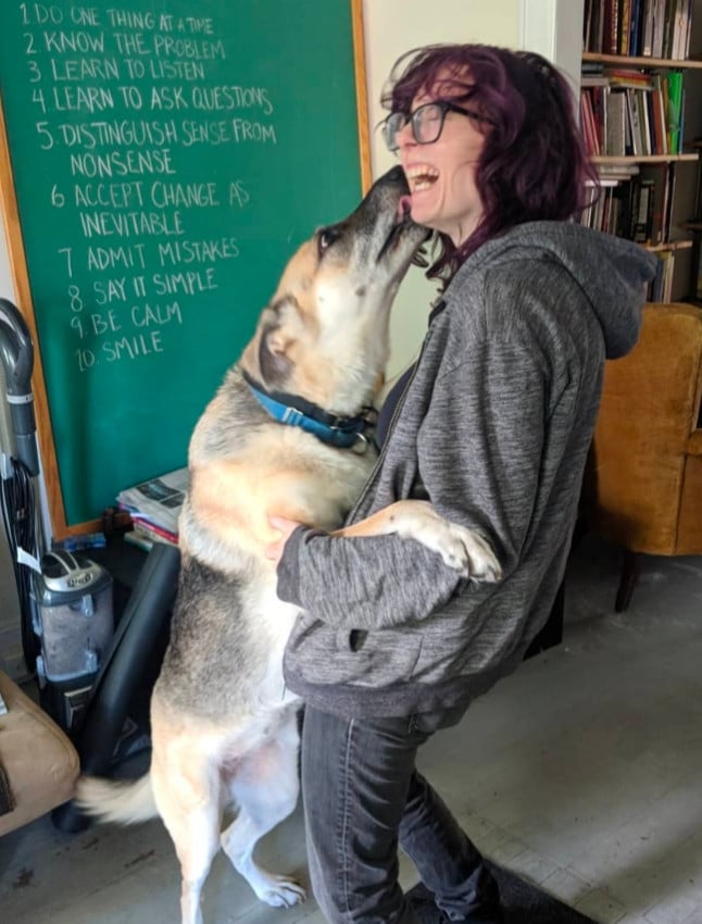 A blonde German Shepherd dog standing on her hind legs to give Ingrid a little smooch on the cheek. Ingrid has purple hair and is holding the dog up in her arms