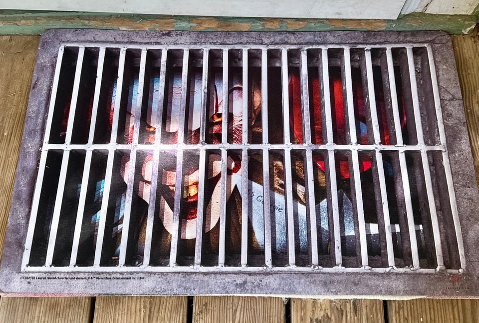 A doormat made to look like a sewer grate, with Pennywise's scary smiling clown face and red hair underneath so it appears as though he's lurking beneath the grate and looking out at whoever is standing on the mat