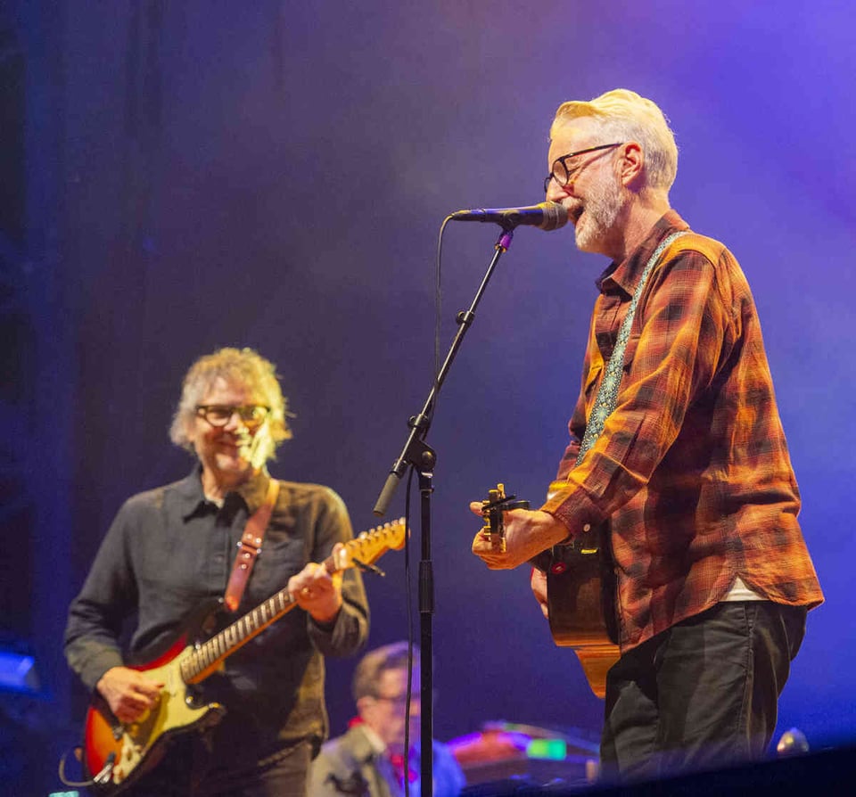 A man with shaggy, graying hair and glasses plays a tobacco sunburst Fender Stratocaster guitar to the right of a man with short white hair and a close-cropped white beard wearing an orange and black plaid shirt who is playing an acoustic guitar and singing into a microphone