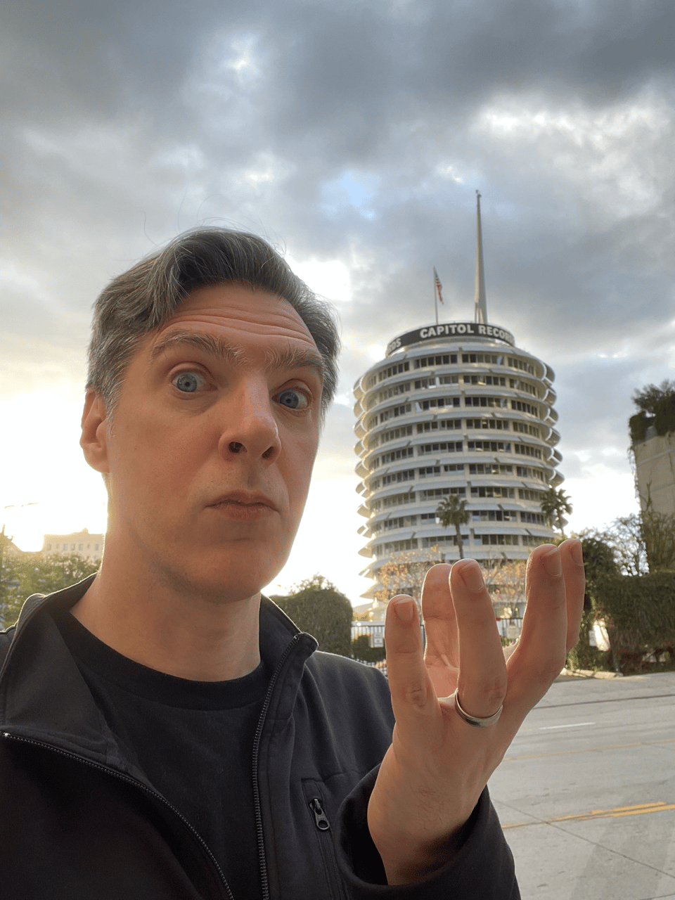 A man looks at the camera and holds a hand up appearing to grab an entire building