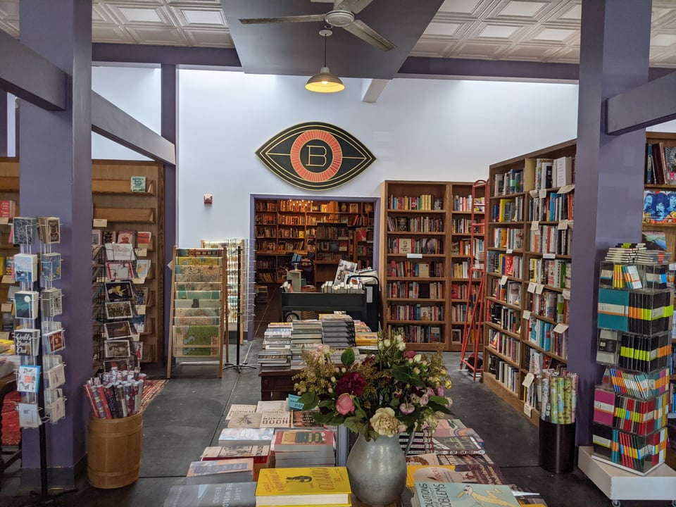 A picture of the Booksmith, with tables full of books out front including a vase full of lovely flowers. Tons of bookshelves line the space plus there's a spinny rack with notebooks and a bin full of wrapping paper. Further back is a door way with an eye over it, with a capital B at the center of the eye
