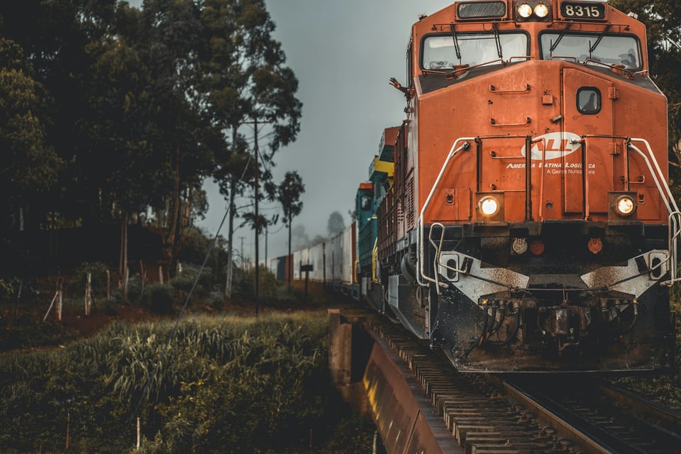 Description: A head-on photo of a red-orange locomotive engine. A trail of attached rail cars is visible behind it. There are trees to one side of the tracks. Photo by Guilherme Stecanella on Unsplash.