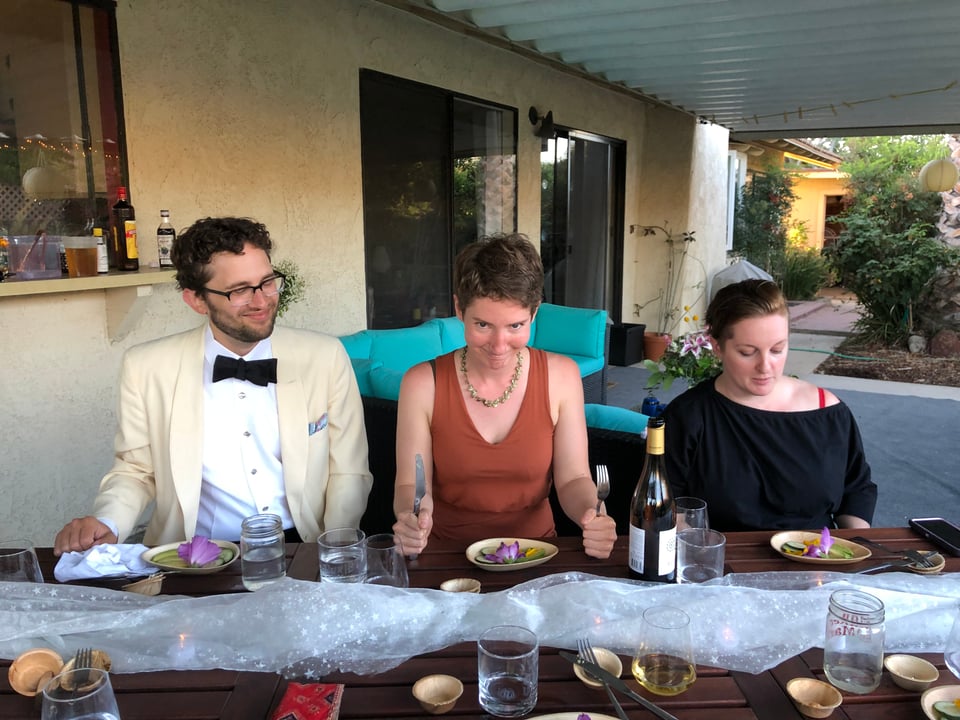 Lucy at a dinner table clutching a knife and fork and glowering at the camera. She's flanked by Gailey and James.