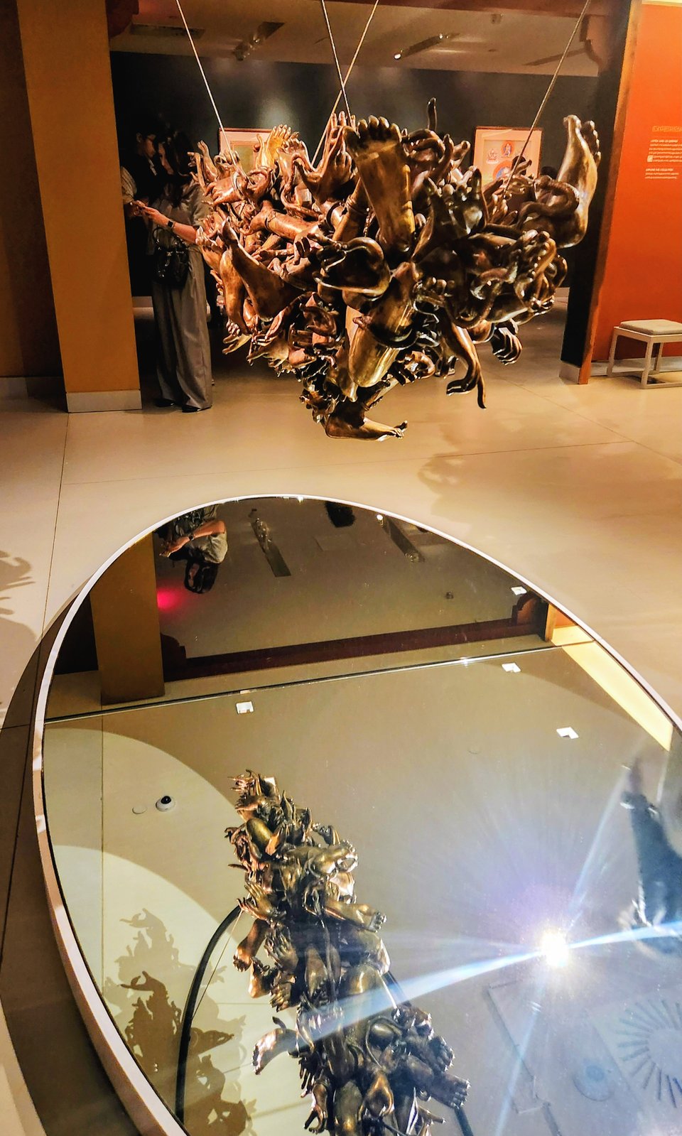 An artwork called "Muted Expressions" rendered in bronze, with various appendages and personal belongings mashed up together, hanging above a large mirror through which you can see the bottom of the work
