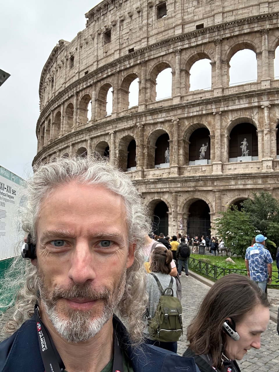 Selfie of white man in navy jacket with an earpiece in, outside the Colosseum