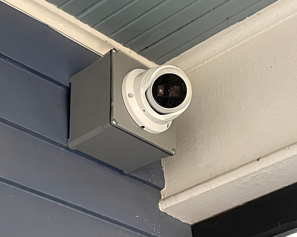 A closeup image of a security camera attached to a metal box, mounted on the exterior of a house with dark blue clapboard siding and white trim.