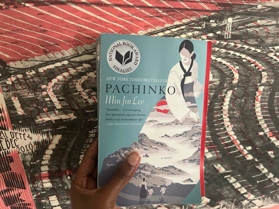 a brown hand holds a blue book titled "Pachinko" by Min Jin Lee against a red backdrop.
