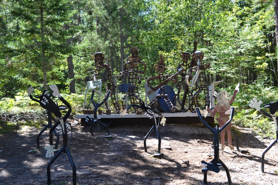 A mteal sculpture of a band performing on a stage, complete with dancing audience, in a wood.