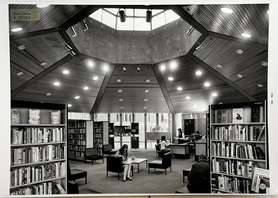 Black and white photo of an octagonal interior space. The open plan space has racks of shelving and some squared off metal frame chairs where a young woman is sitting reading. But the real focus is of the wood-lined ceiling and the octagonal concrete ring that holds it in place. Above the ring is an equally octagonal lantern that is letting in light.
