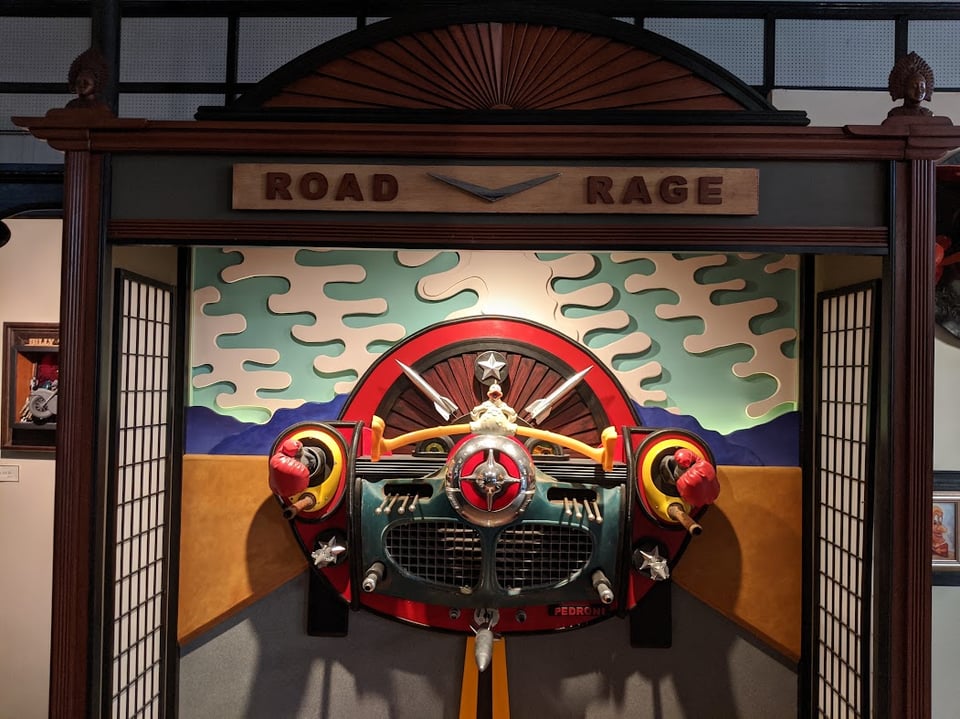 A sign says ROAD RAGE. There is a chicken driving an old-fashioned car with boxing gloves coming out of the headlights.