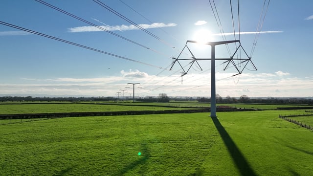 Photo of a T shaped pylon in the Englush countryside. The sky is blue and lightly cloudy, and the grass is bright green.