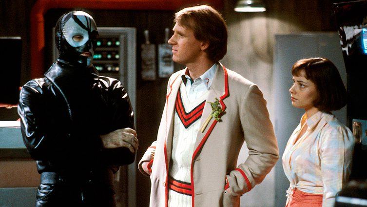 The 5th Doctor and Peri face to face with Sharaz Jek, in a menacing black and white leather mask, with one scarred hand visible.