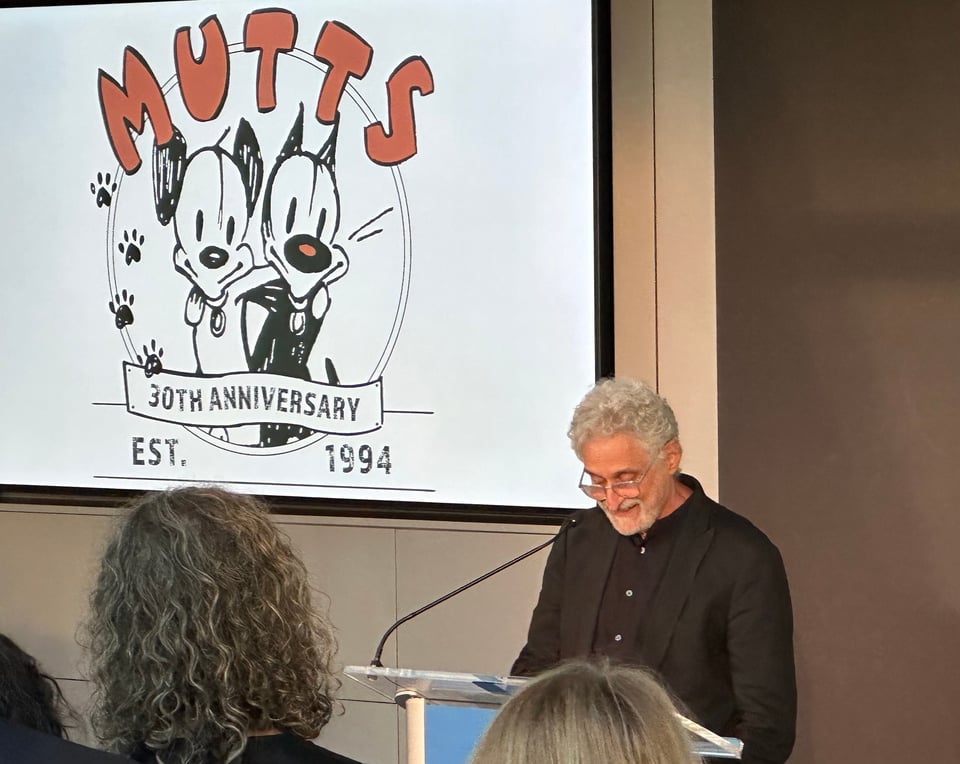 Photo of cartoonist Patrick McDonnell standing at a lectern giving a speech, in front of a screen showing the logo for the 30th anniversary of the Mutts comic strip, which has Earl and Mooch with arms over each other's shoulders