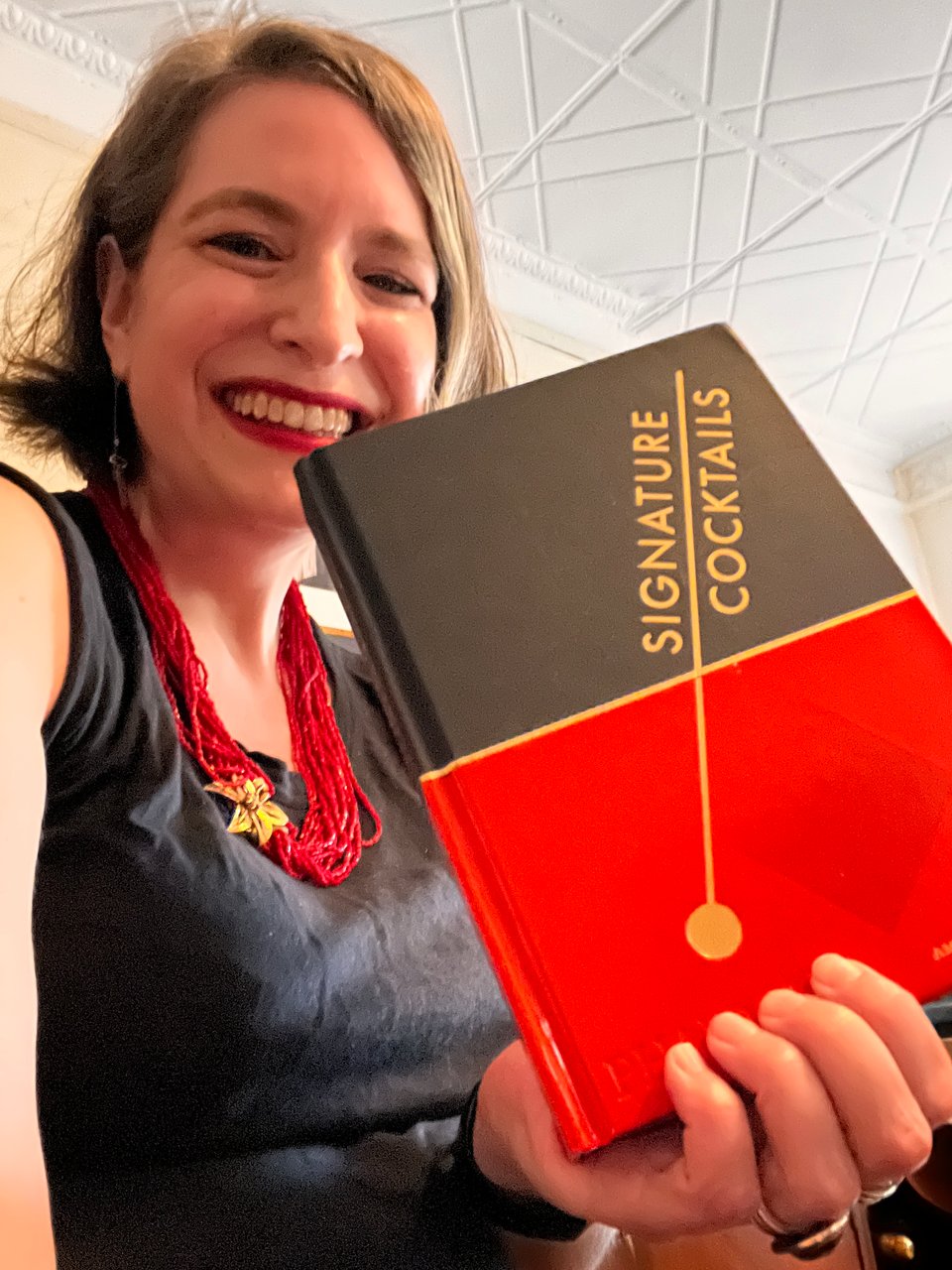 slightly disheveled author Amanda Schuster holds copy of new book, Signature Cocktails, 2023
