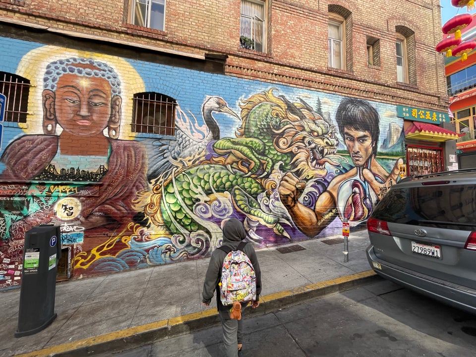 A boy in a grey hoodie walks towards a mural in ChinaTown. The mural has Buddha, Chinese dragon, crane, Bruce Lee and other Asian elements.