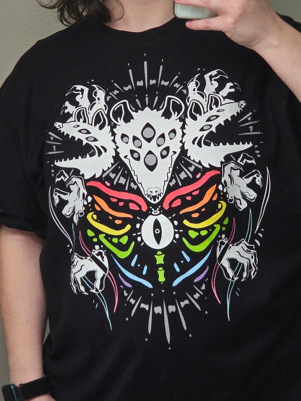 Mirror selfie of someone wearing the biblically accurate possum shirt in pride gay ass rainbow. Its a black and white design of a three headed possum with multiple eyes and rainbow ribs.