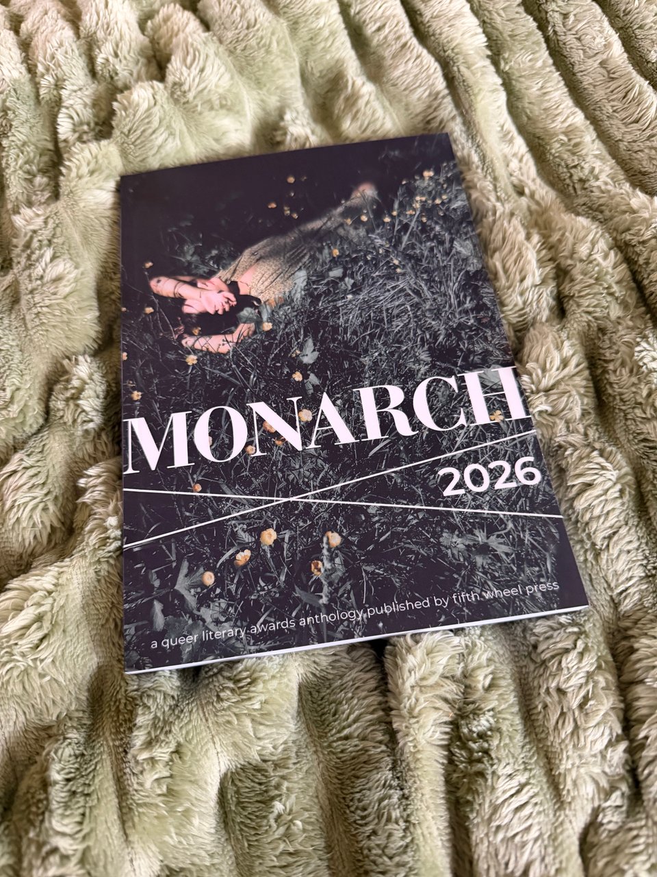 Picture of a book laying on a soft, fuzzy sage-green blanket. The title reads: MONARCH 2026, a queer literary awards anthology published by fifth wheel press. A white person in a floral dress is laying with an arm over their face in a field of dark green grass and foliage, with yellow flowers.
