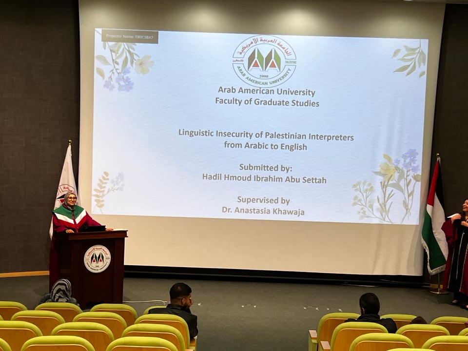 A woman on stage at a podium with a large screen showing a title slide:  Arab American University Faculty of Graduate Studies, Linguistic Insecurity of Palestinian Interpreters from Arabic to English, Submitted by Hadil Hmoud Ibrahim Abu Settah, Supervised by Dr. Anastasia Khawaja