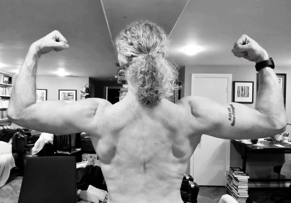 b/w photo of back of white man with long hair, flexing biceps and back. He looks kinda ripped, and you'd be surprised that he's in his 50s.