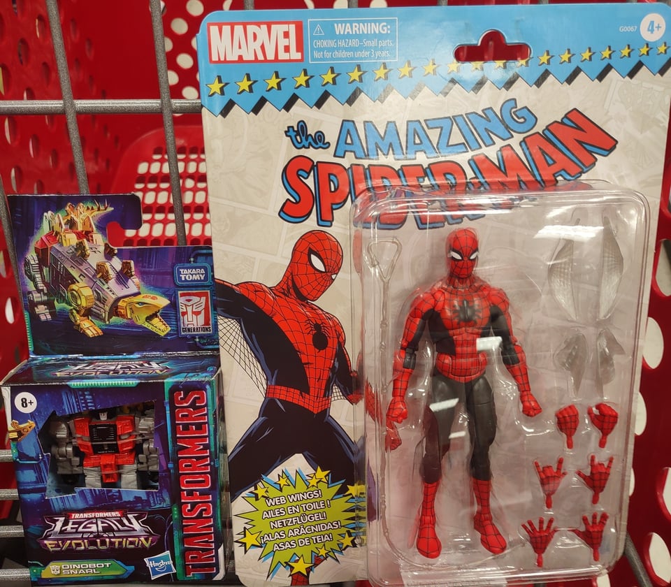 Two packaged toys sit in the "seat" of a shopping cart--a small Transformers robot-dinosaur and a Marvel Amazing Spider-Man in a red and black costume.