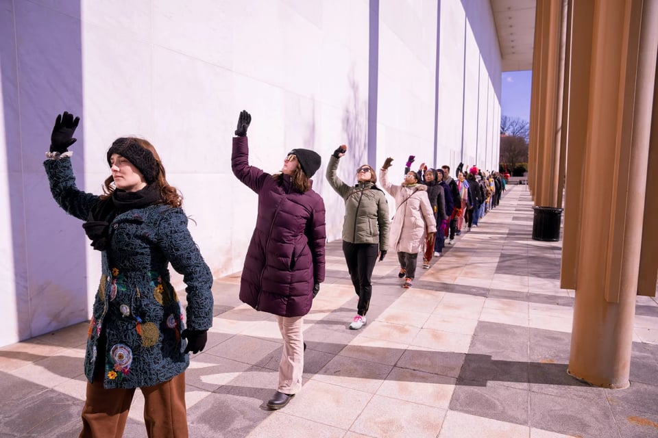 A line of dancers raise their arms as part of a choreography outside the Kennedy Center on a cold day.