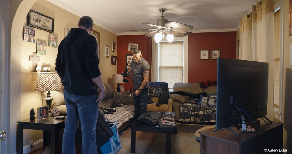 Image of two men in a living room. The couch is covered in various types of guns. Image is by Auberi Edler.