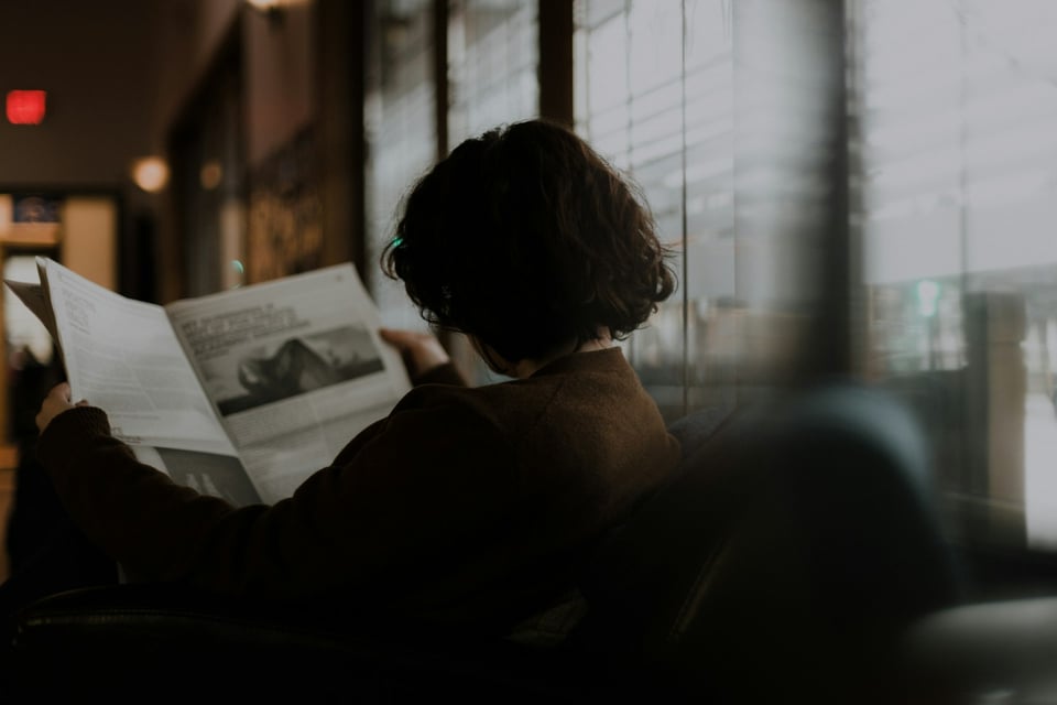 A dim, slightly blurred photo of a woman reading a newspaper.  Photo by Priscilla Du Preez 🇨🇦 on Unsplash.