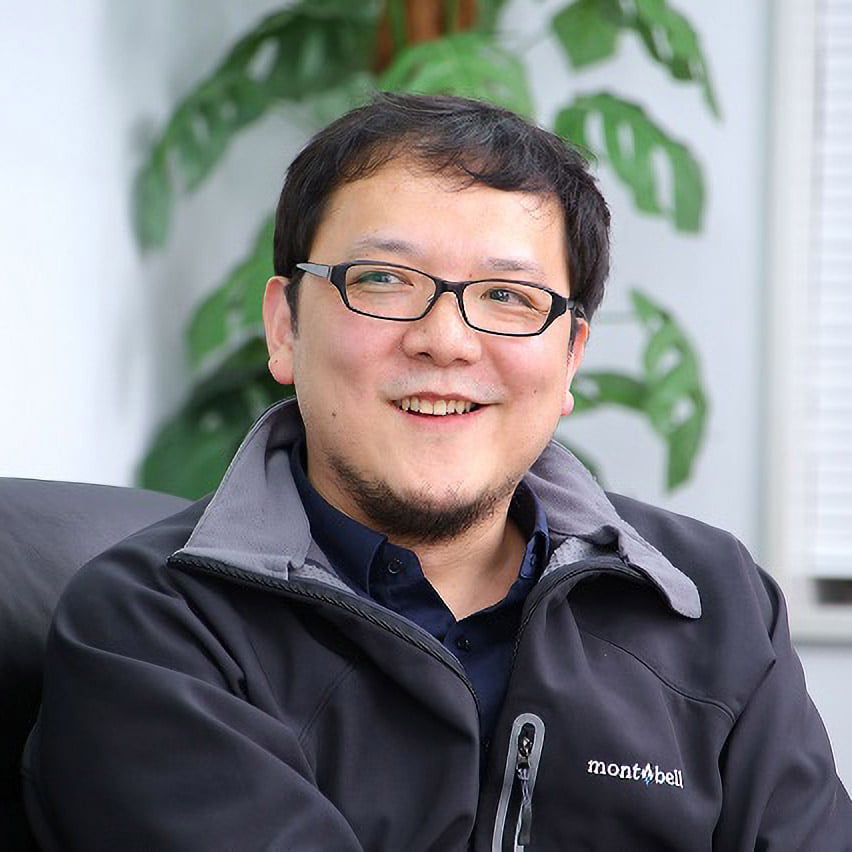 A picture of Japanese game designer Hidetaka Miyazaki wearing a black Montbell pullover jacket and black glasses, smiling to the camera