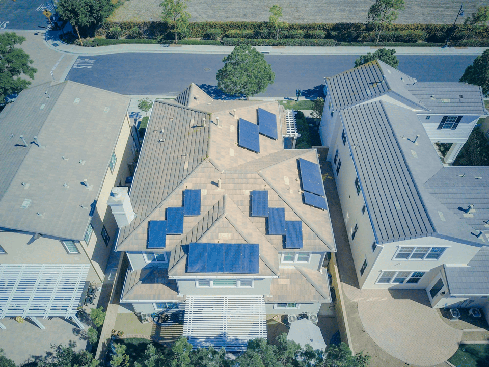 An aerial image taken of three houses, shot from perhaps 100 feet above them. They are suburban two-story houses, and the middle one has perhaps a dozen solar panels scattered around the slanted roof. It is a sunny day, and the picture has a very cheery field to it
